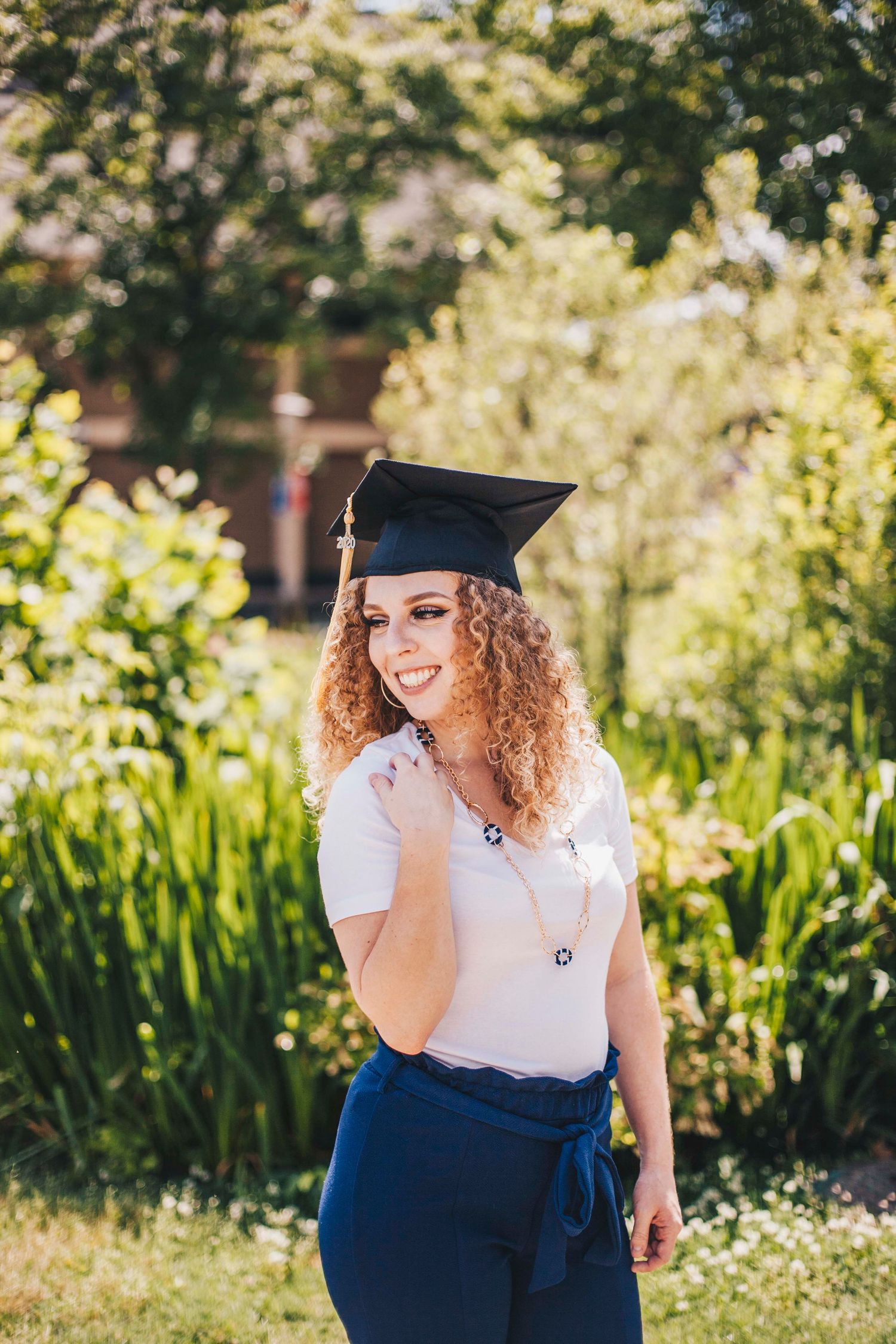 A smiling curly-haired graduate wearing a black mortarboard cap and blue jeans poses outdoors.