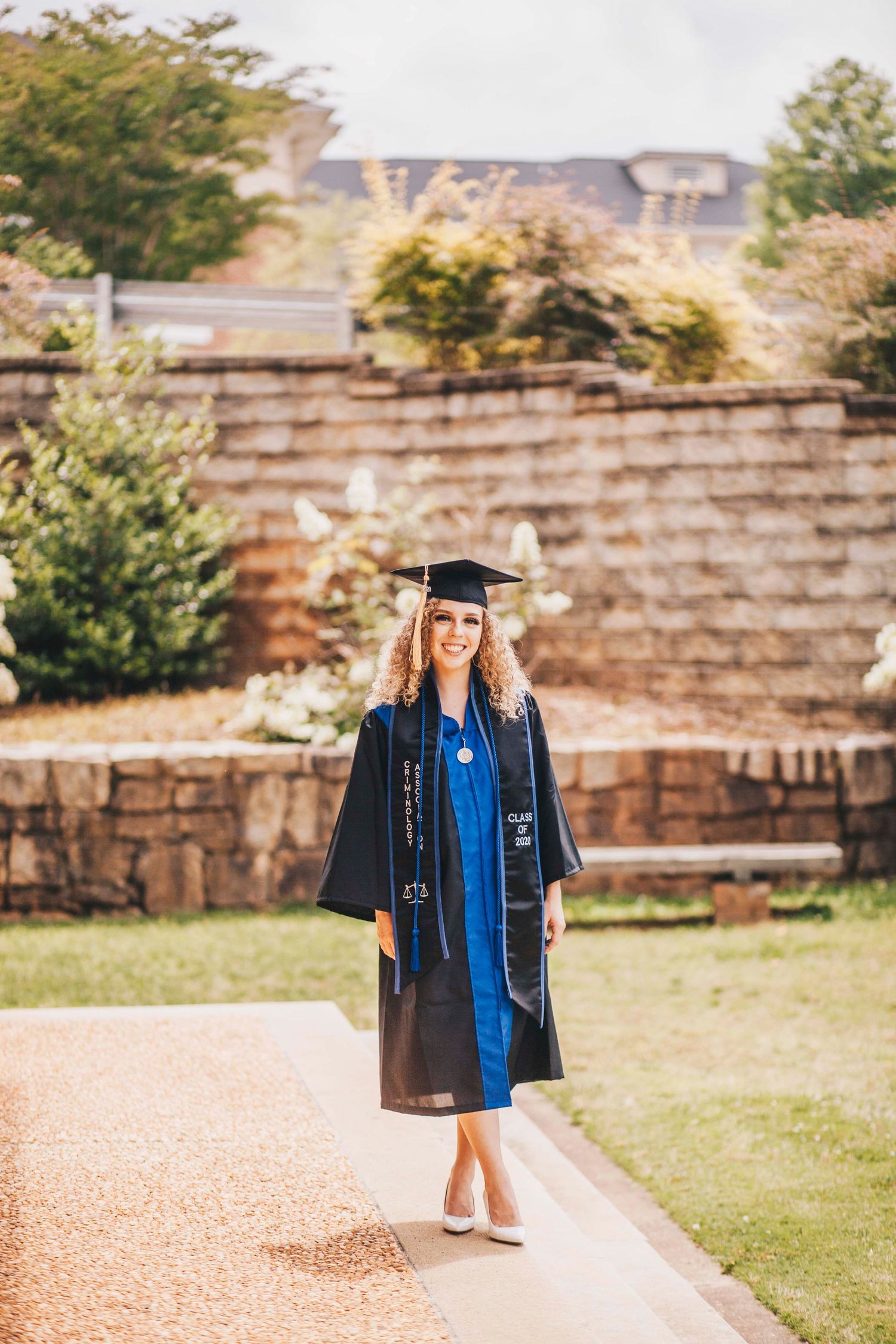 Smiling female graduate in black gown and blue stole on campus path.