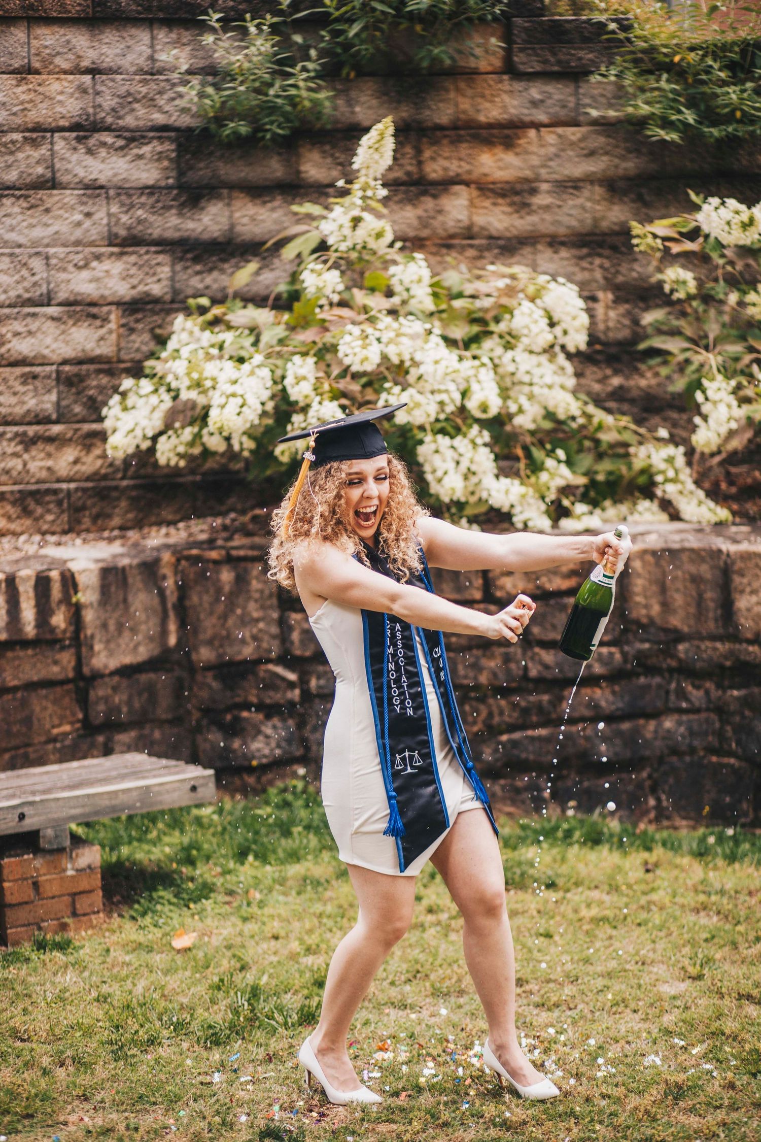 Excited graduate in cap and sash pops a champagne bottle outdoors near a stone wall and flowers.