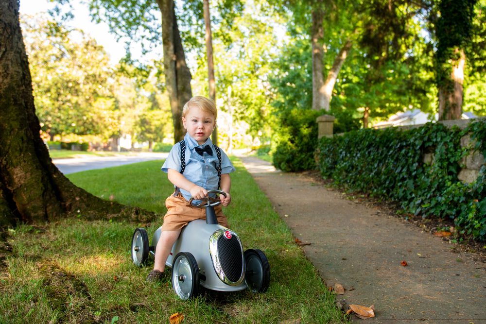 Child riding pedal car along tree-lined path on autumn day.