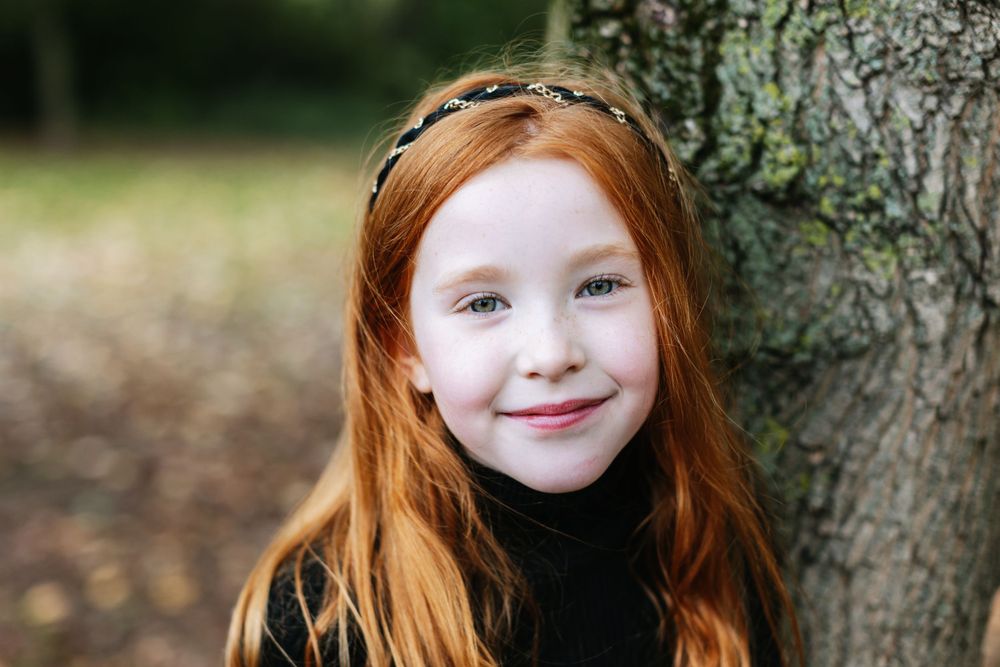 A portrait of a young person with red hair leaning against a tree trunk.
