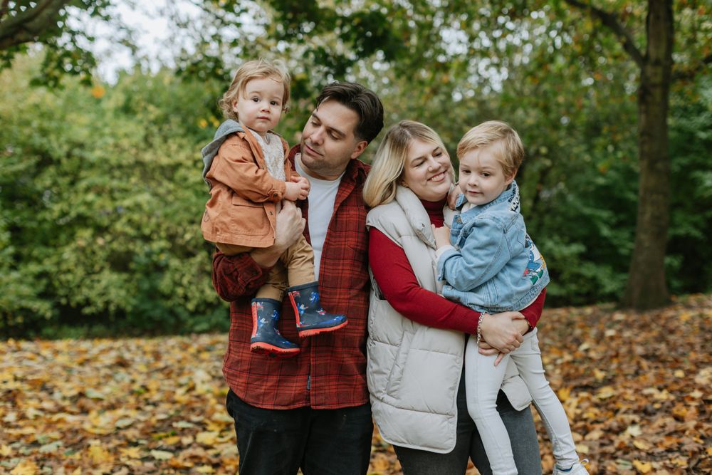 Family of four posing together among fallen autumn leaves in wooded setting.