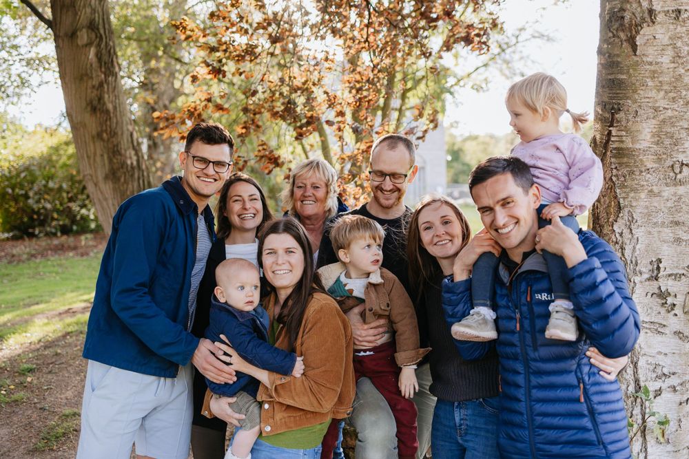 A large family group poses together outdoors against a backdrop of autumn trees.