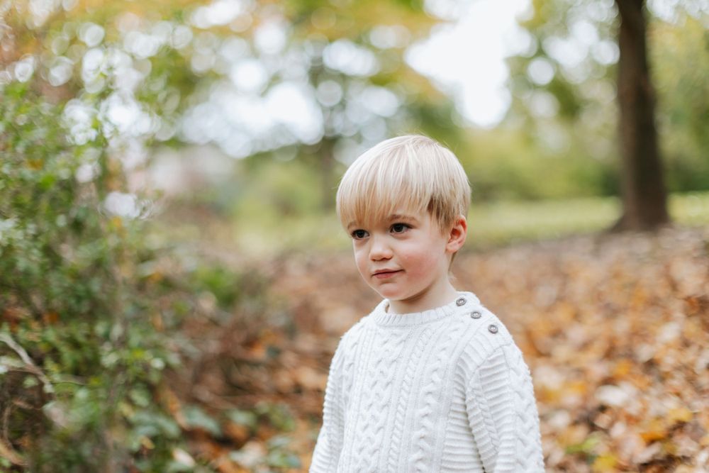 A child in a white cable knit sweater stands among fallen autumn leaves.