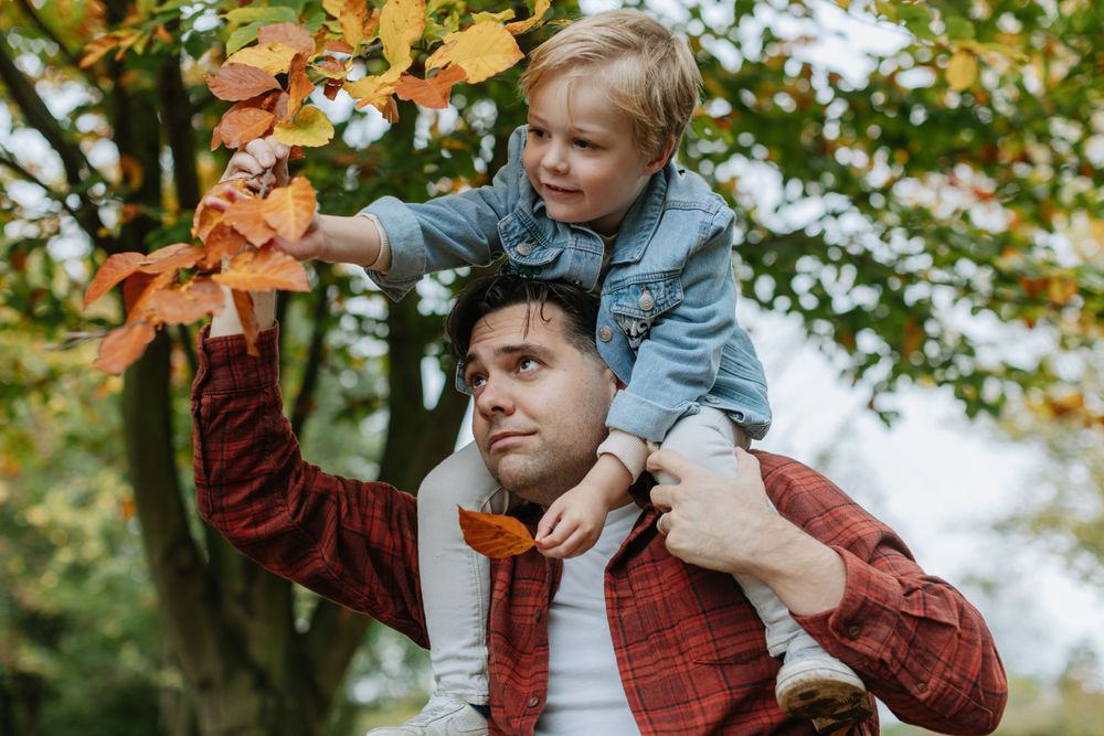 Joyful autumn moment with child riding on shoulders reaching for fall leaves.