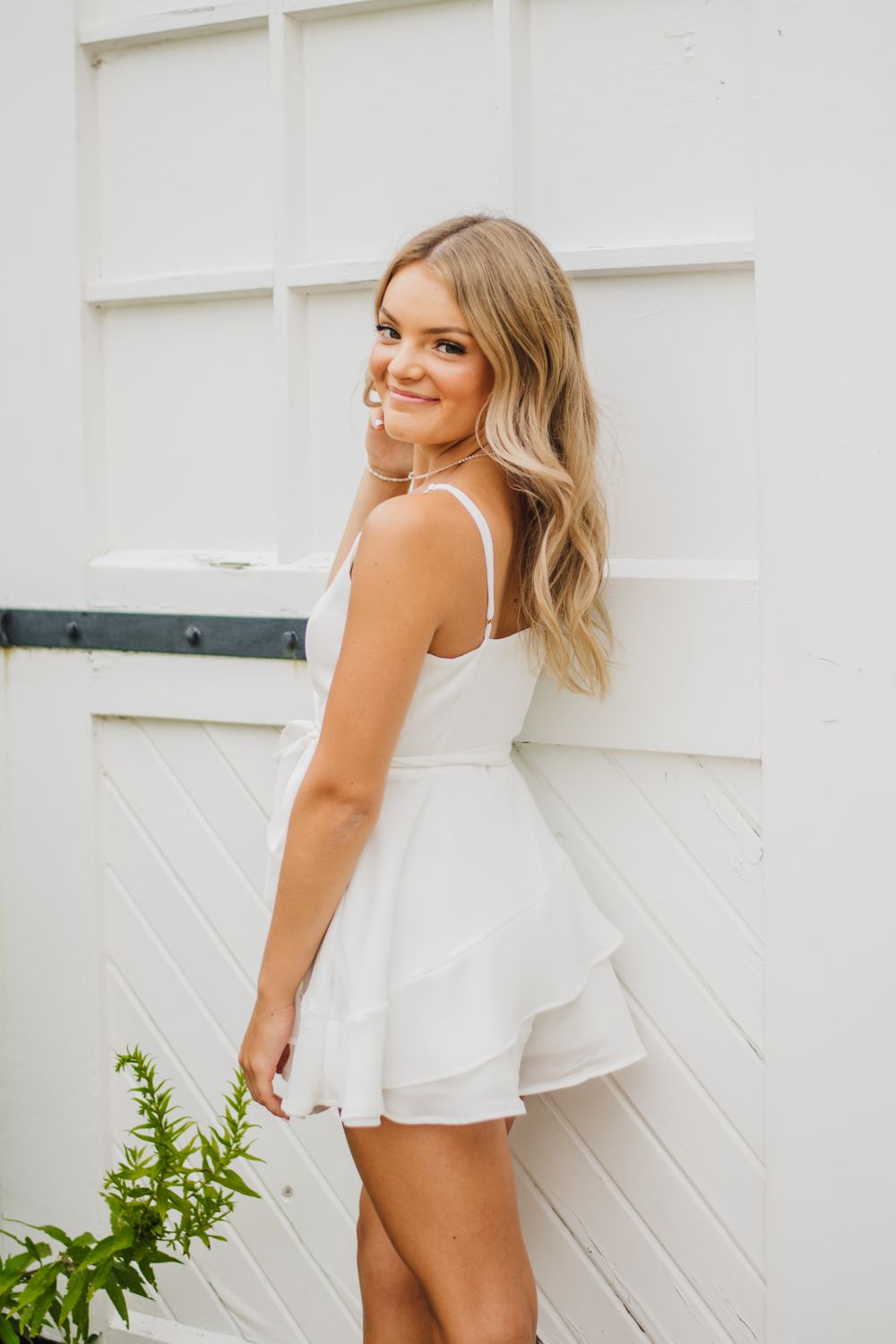 White ruffled romper photographed against a white wall with green foliage.