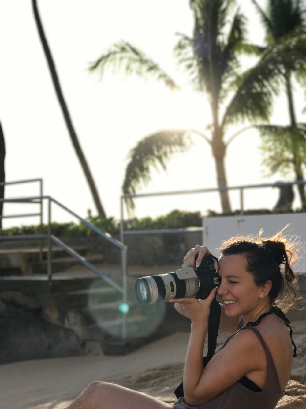 Photographer with telephoto lens captures images at sunset among palm trees.