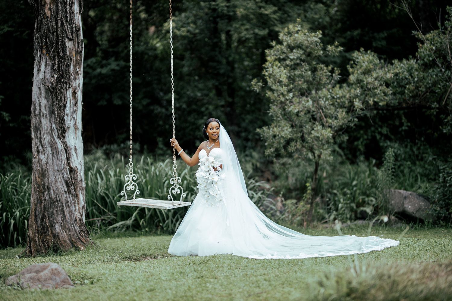 Bride in flowing white dress sitting on garden swing in natural setting.