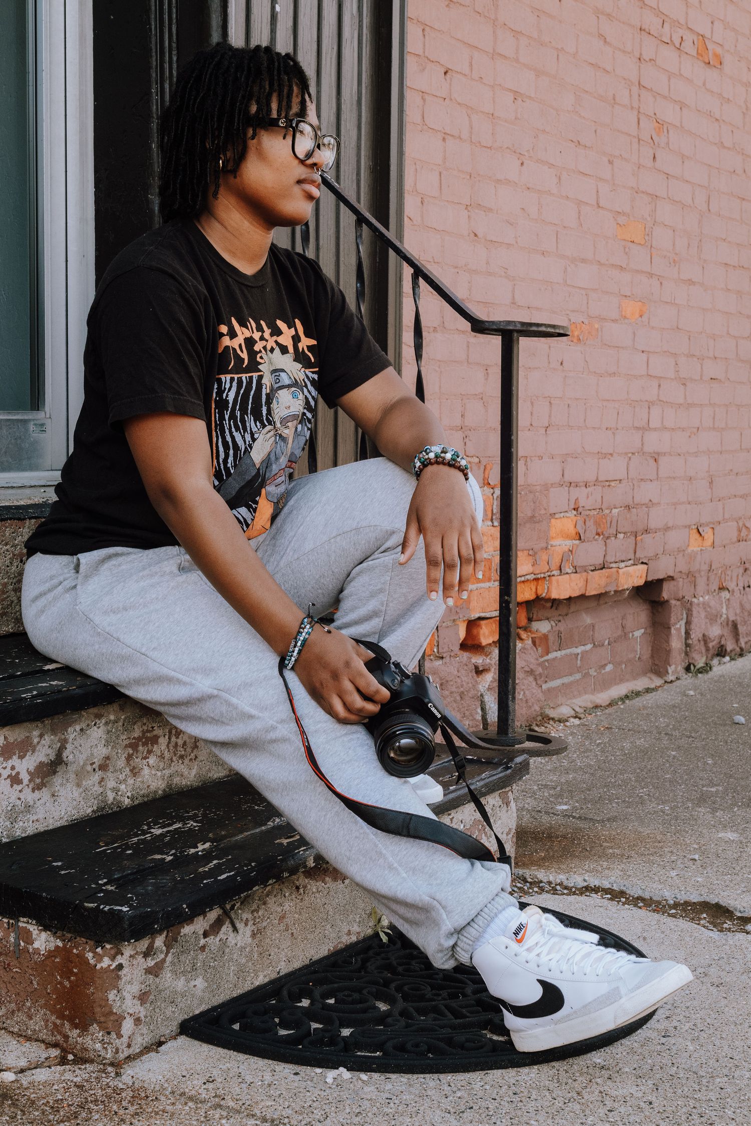Photographer with dreadlocks and glasses sits on steps with camera wearing black t-shirt and grey pants.