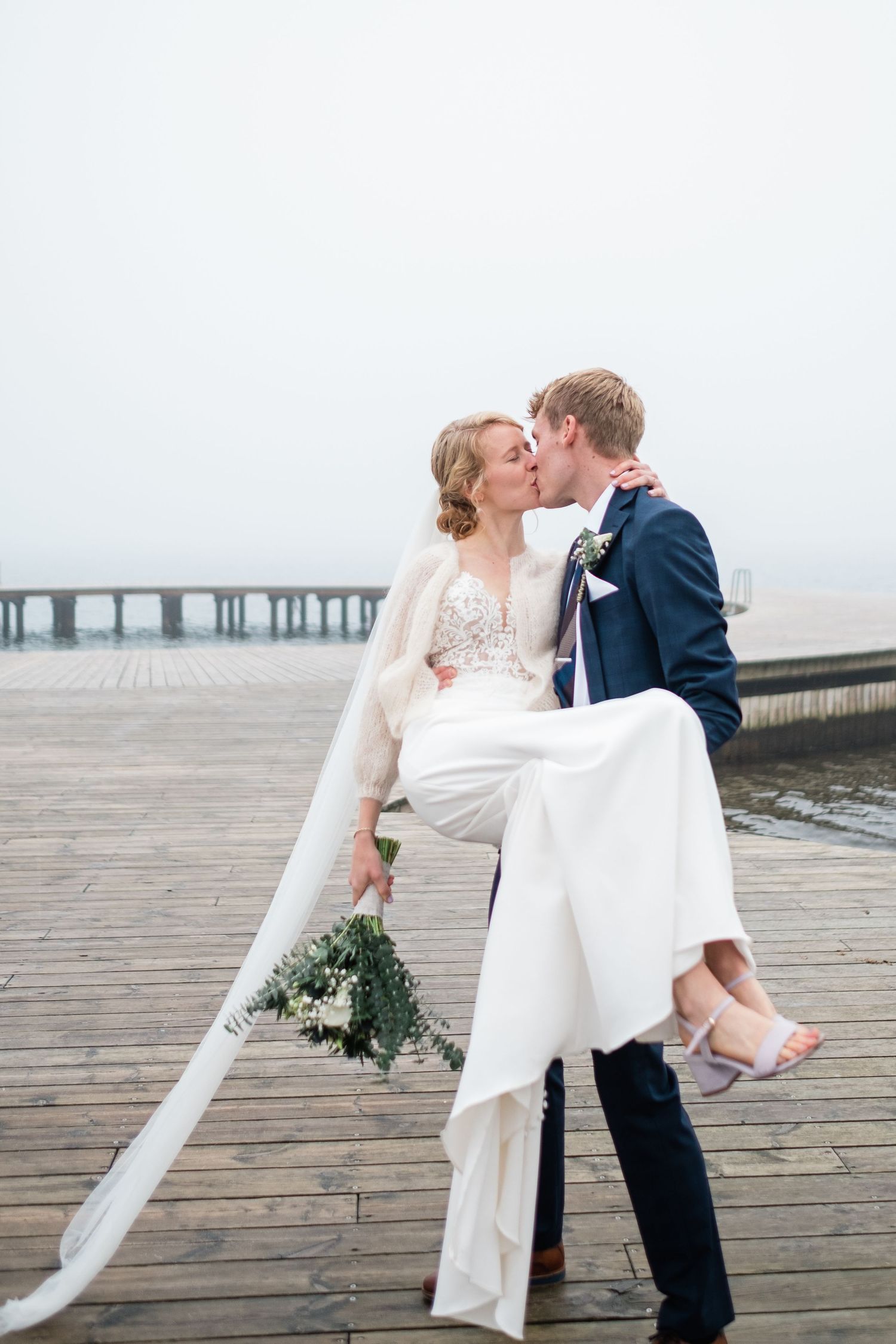 Groom lifts bride in romantic pose on wooden pier by the ocean.
