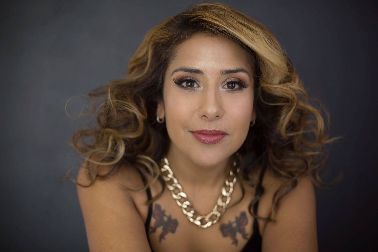 Close-up portrait of a woman with curly blonde hair and chest tattoos wearing a gold chain necklace.