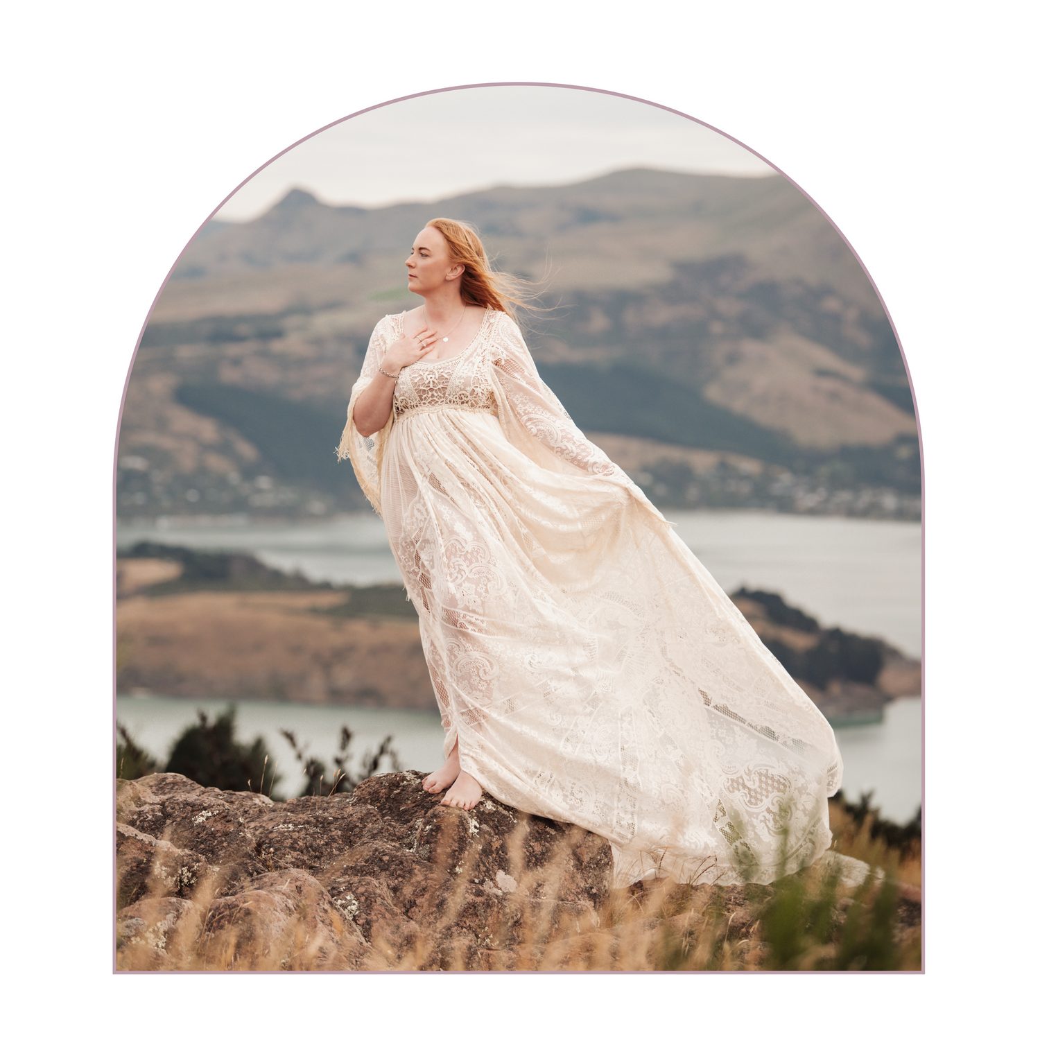 Dramatic maternity portrait in flowing cream dress on cliff overlooking mountain lake.