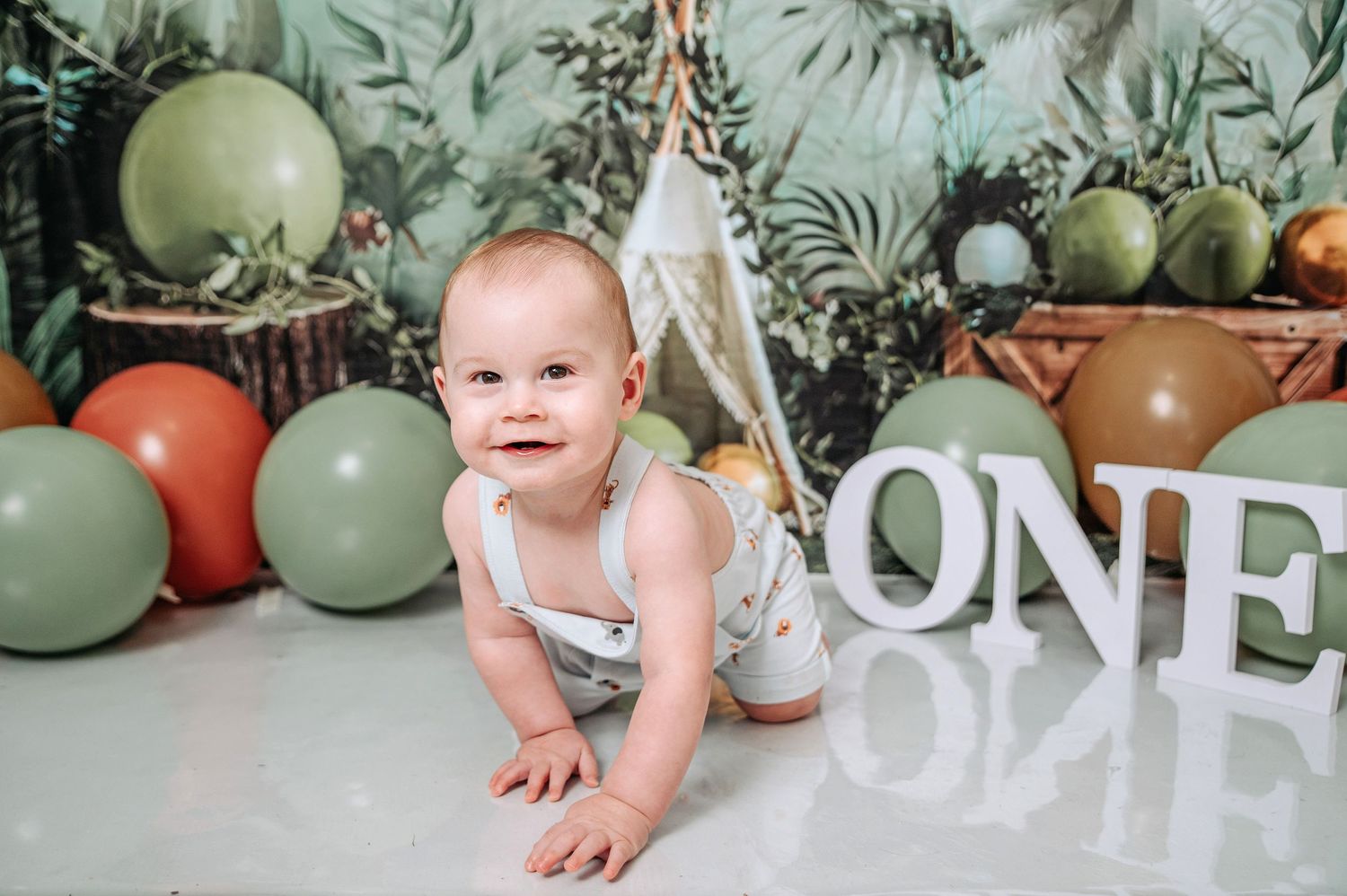 First birthday photoshoot with tropical backdrop, white teepee, balloons and decorative 'ONE' letters.