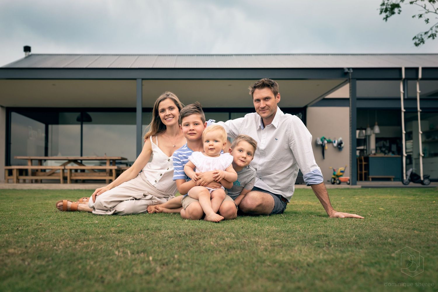 Family relaxing on grass lawn in front of modern house with picnic shelter.