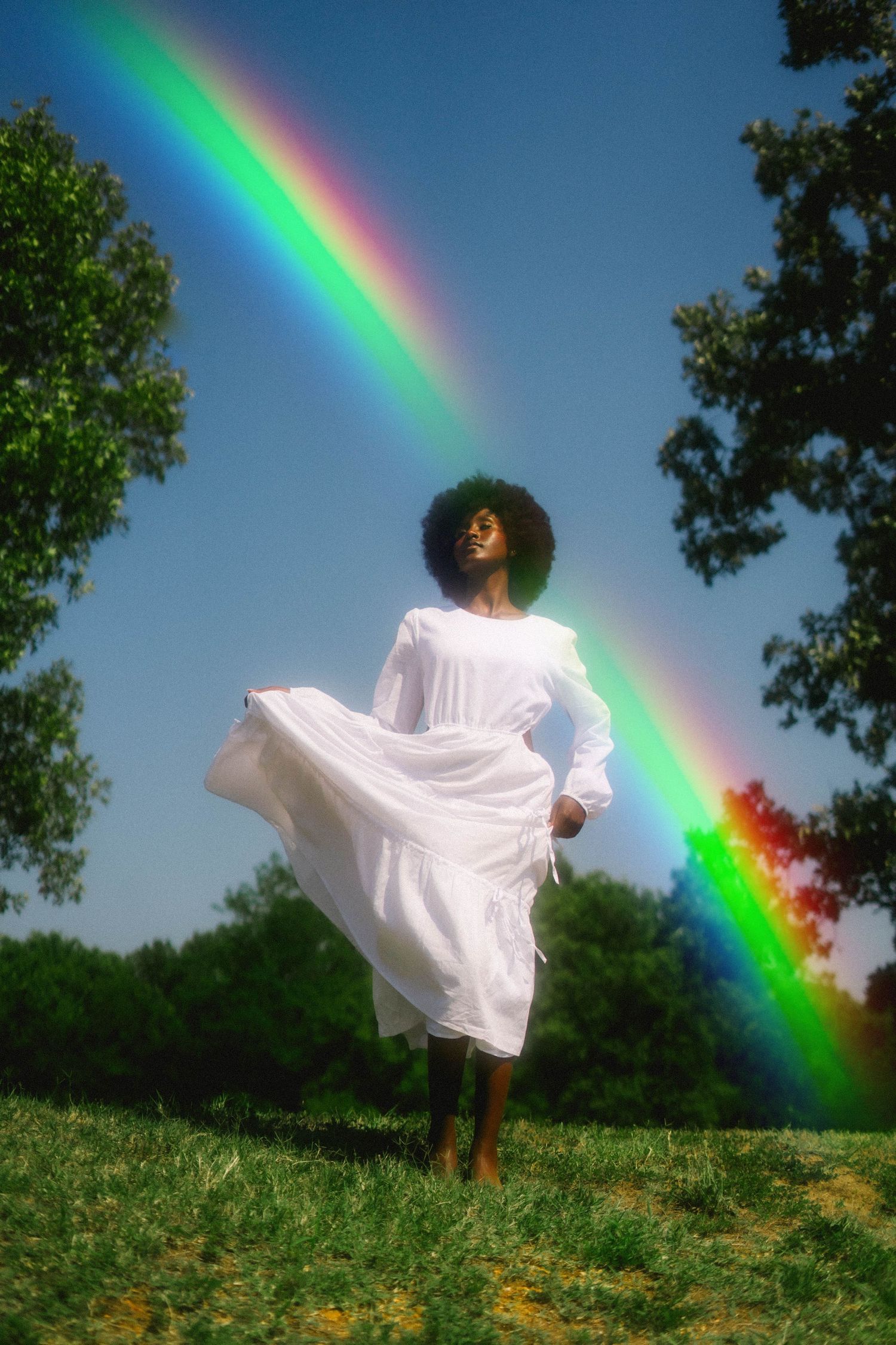 Person in flowing white dress dancing in field with vibrant rainbow in background.