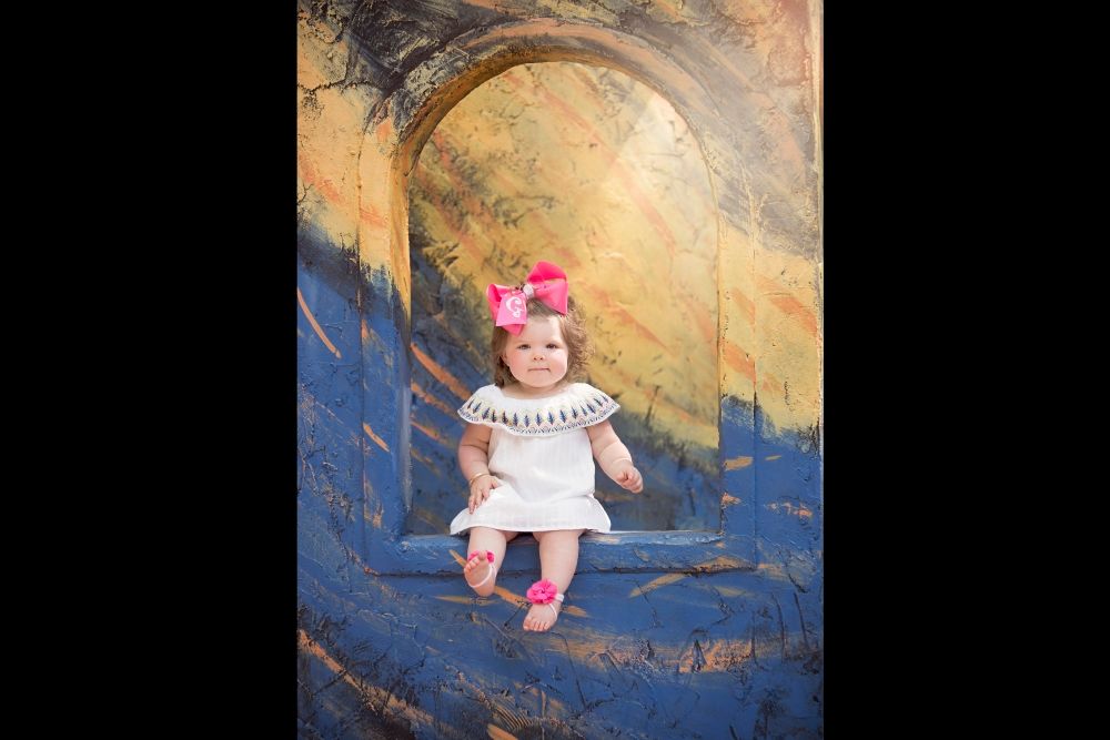 A small child sitting in a colorful portrait backdrop wearing a white dress and pink flower hairpiece.