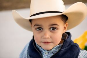 Young child wearing a white cowboy hat and blue jacket against neutral background.