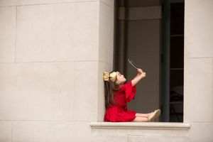 Person in red dress sitting in window ledge against white wall taking selfie.