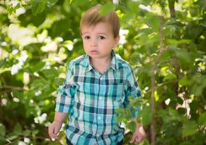 Young child in plaid shirt standing among green foliage outdoors.