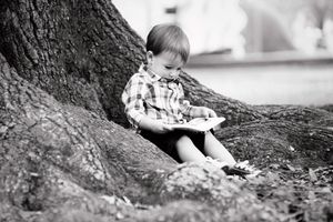 Child in plaid shirt reading while sitting against large tree trunk in black and white.