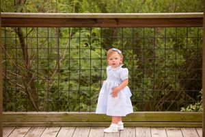 Child in light blue dress standing on wooden deck with green foliage background.