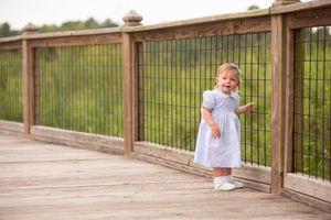 Small child in white dress stands on wooden boardwalk with railing on sunny day.