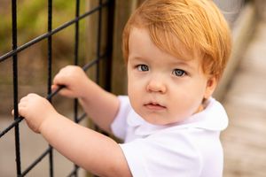 Young child in white shirt holds onto metal fence while looking to side.