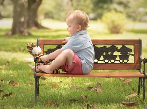 Small child sits on decorative metal park bench with toy on sunny day.