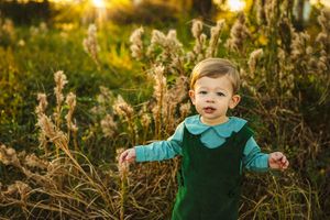 Child in teal outfit stands among tall grass during golden sunset.