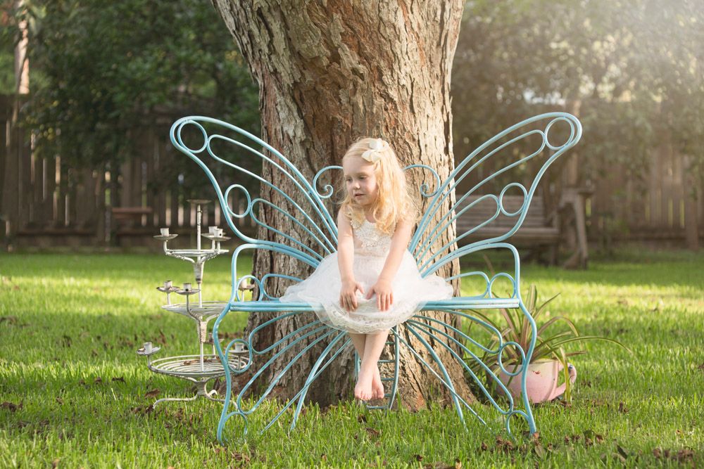 Child in white dress sits on fairy bench with sparkly wings in garden setting.