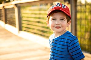 A young one wearing a striped blue shirt and red baseball cap smiles outdoors.