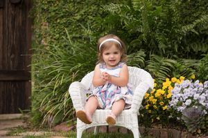 A small figure sits in a white wicker chair among garden flowers.