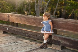 Child in blue dress and boots sitting on wooden bench outdoors.
