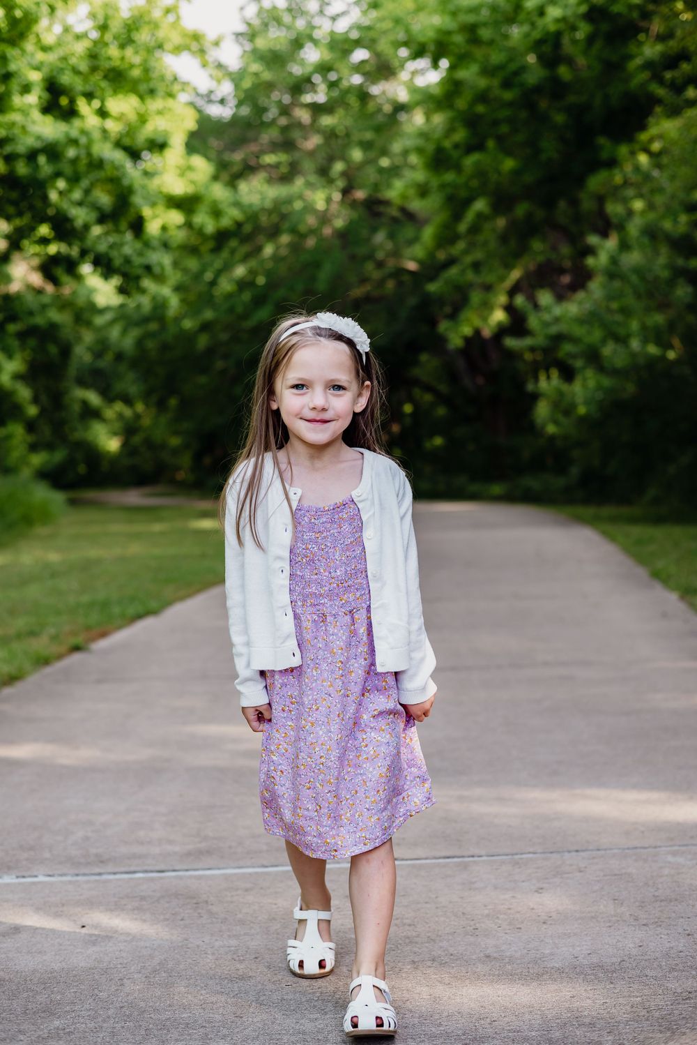 Young child in purple dress and white cardigan walking on paved path.