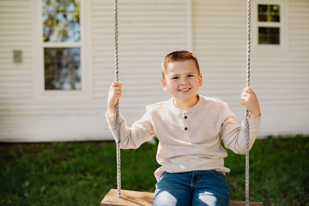 Child swinging on a wooden rope swing outside a white house on a sunny day.