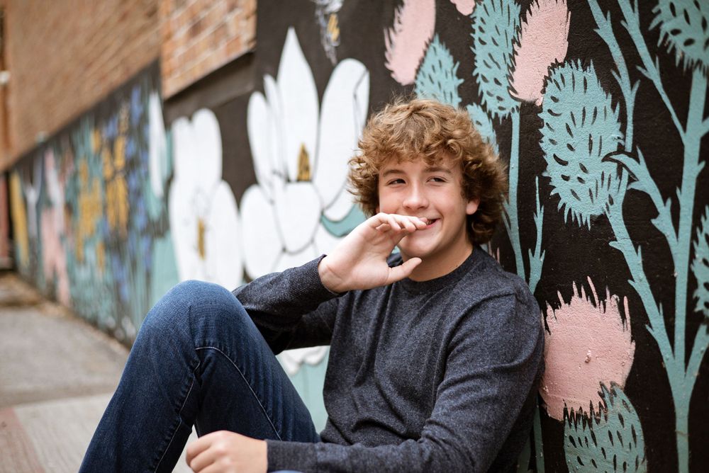 Person with curly hair wearing grey sweater sitting against colorful graffiti wall.