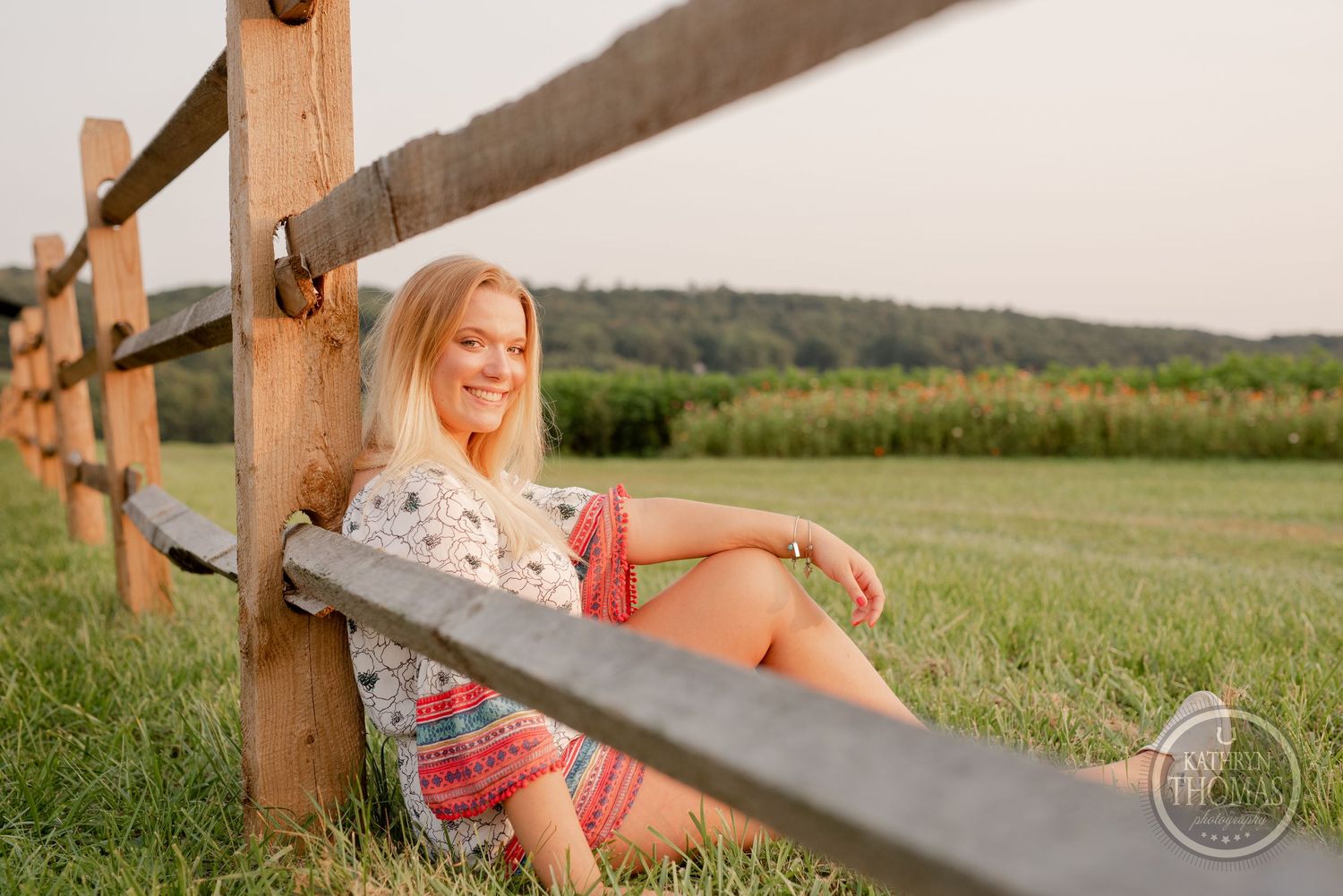 A woman relaxes on a wooden fence in a countryside setting during sunset.
