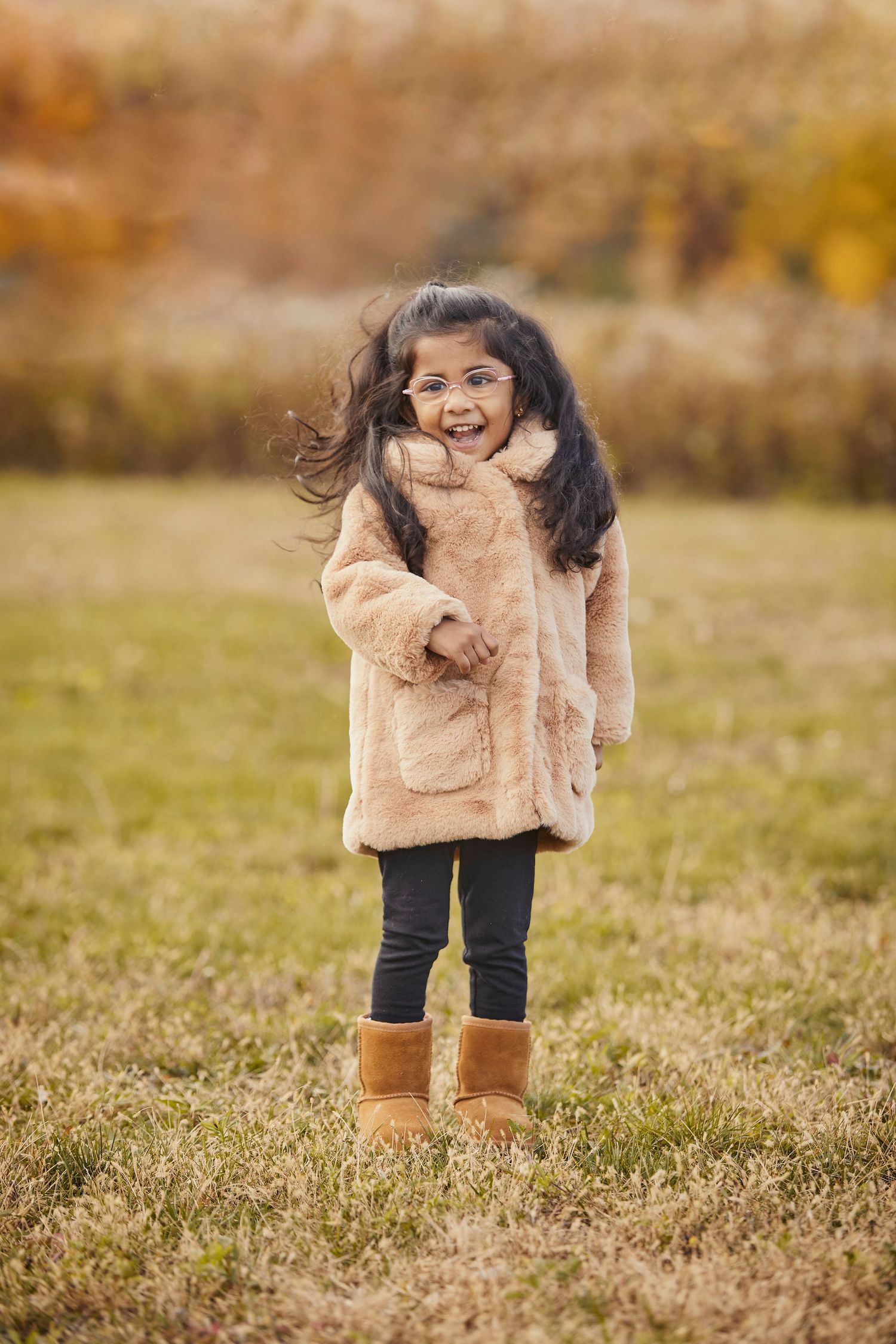 A joyful expression shines through while posing in a cozy beige winter coat and tan boots on autumn grass.