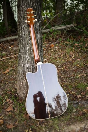Acoustic guitar leaning against tree reflects silhouette of couple in its mirrored surface.