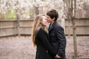 A romantic couple embraces outdoors wearing winter coats among bare trees.