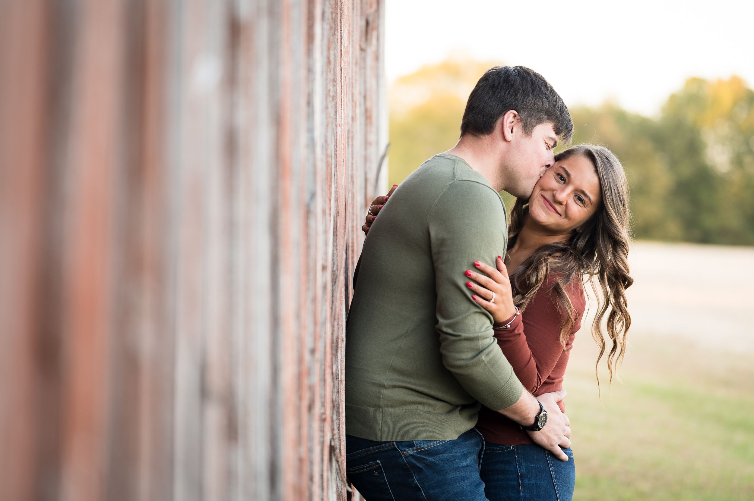 Couple laughing and embracing against rustic barn wall during sunset engagement session.