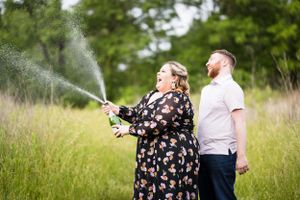 Person sprays champagne outdoors while companion watches in amusement.
