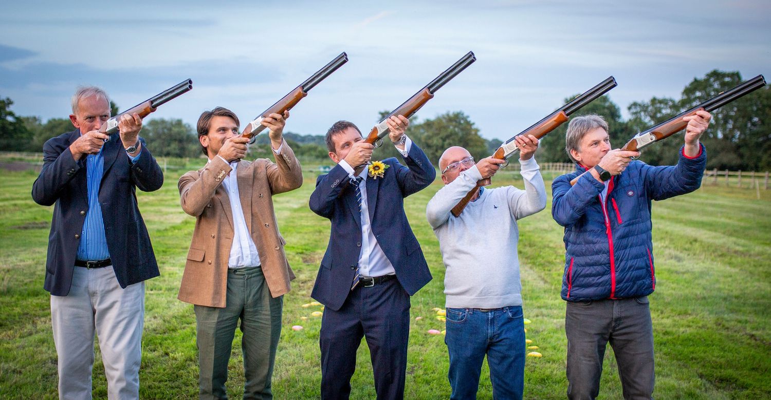 Group photo of wedding guests lined up with clay shooting rifles in countryside setting.