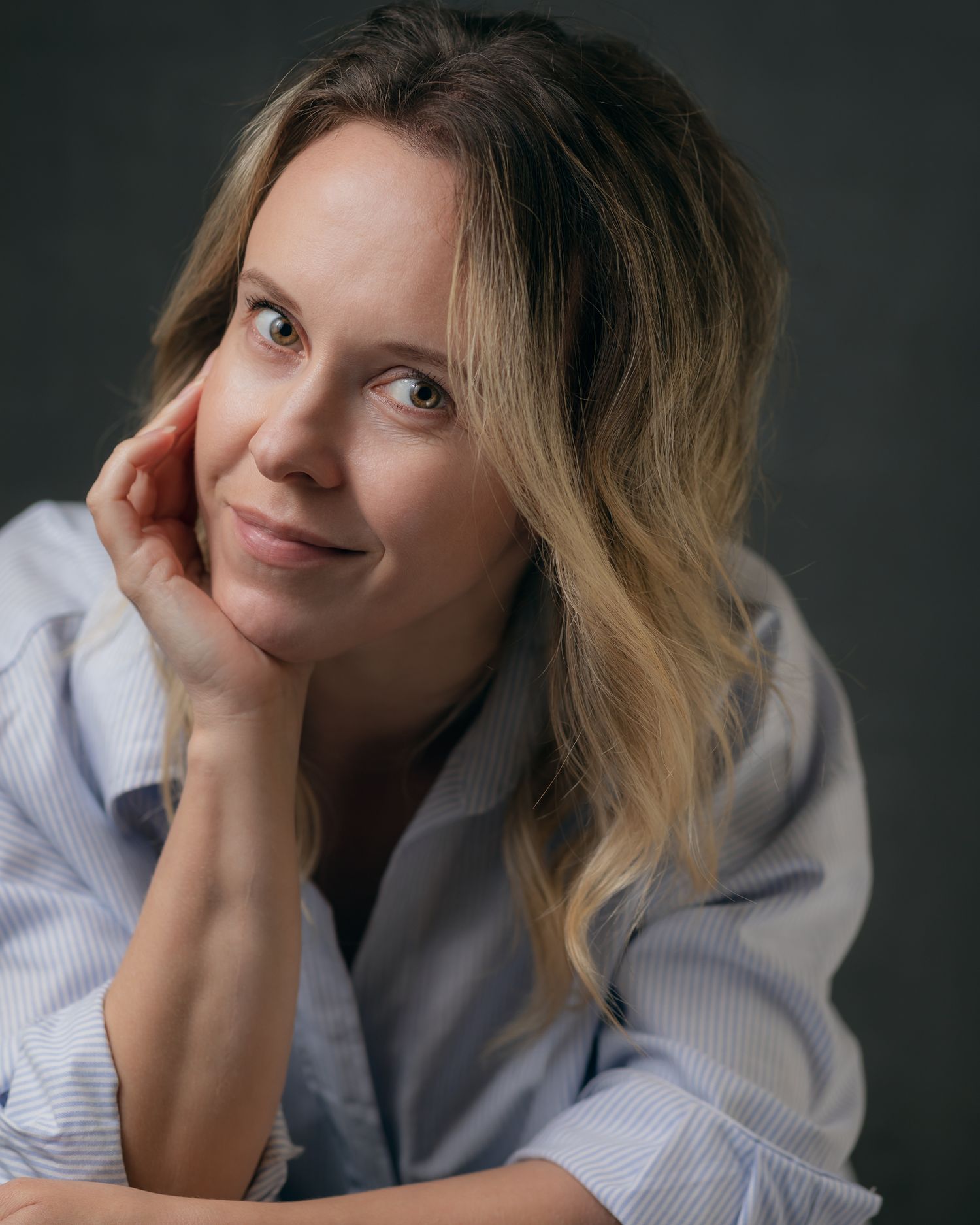 Casual portrait of someone with blonde hair resting head on hand against dark backdrop.