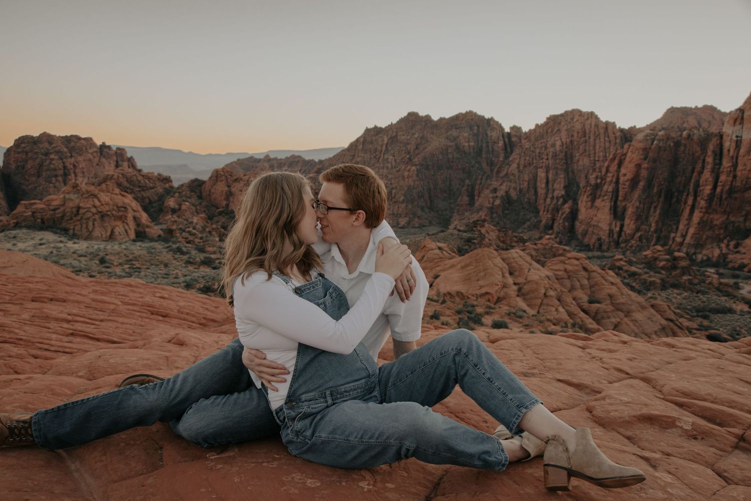 A couple embraces on red rocks during sunset at a desert location.