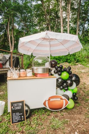 Outdoor party setup with white umbrella, balloons, and football decorations in a wooded setting.