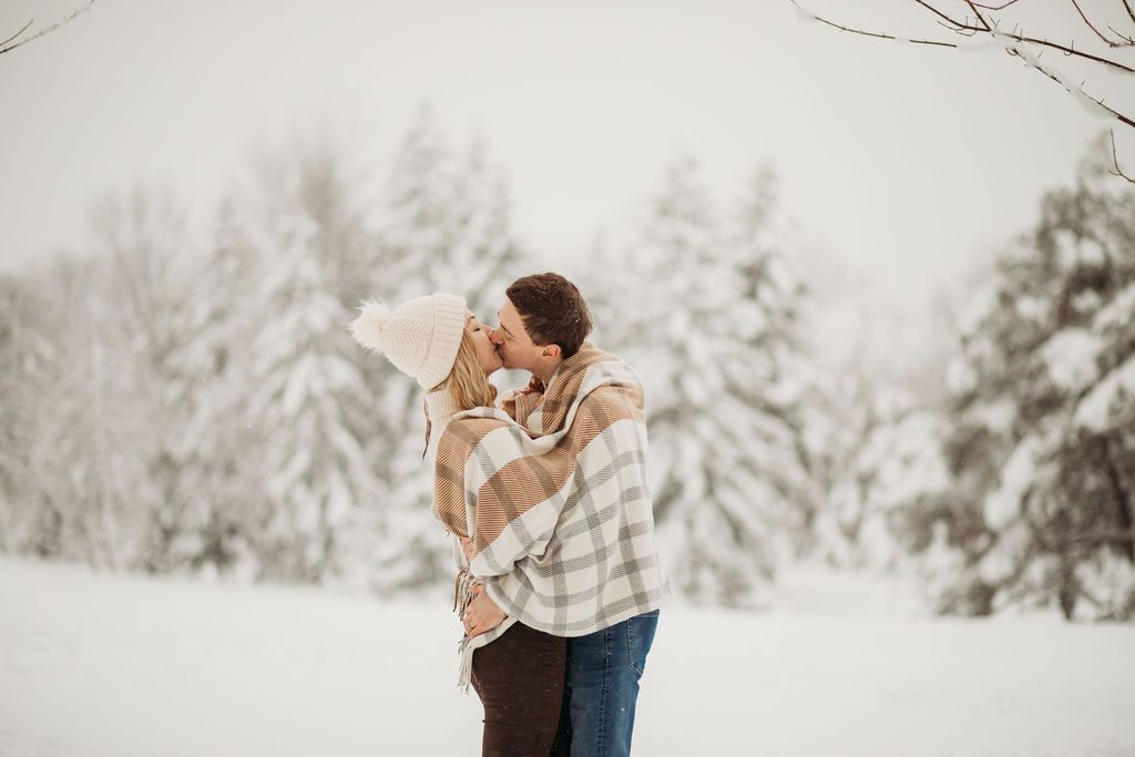 A couple shares a tender embrace while wrapped in a plaid blanket against a winter forest backdrop.