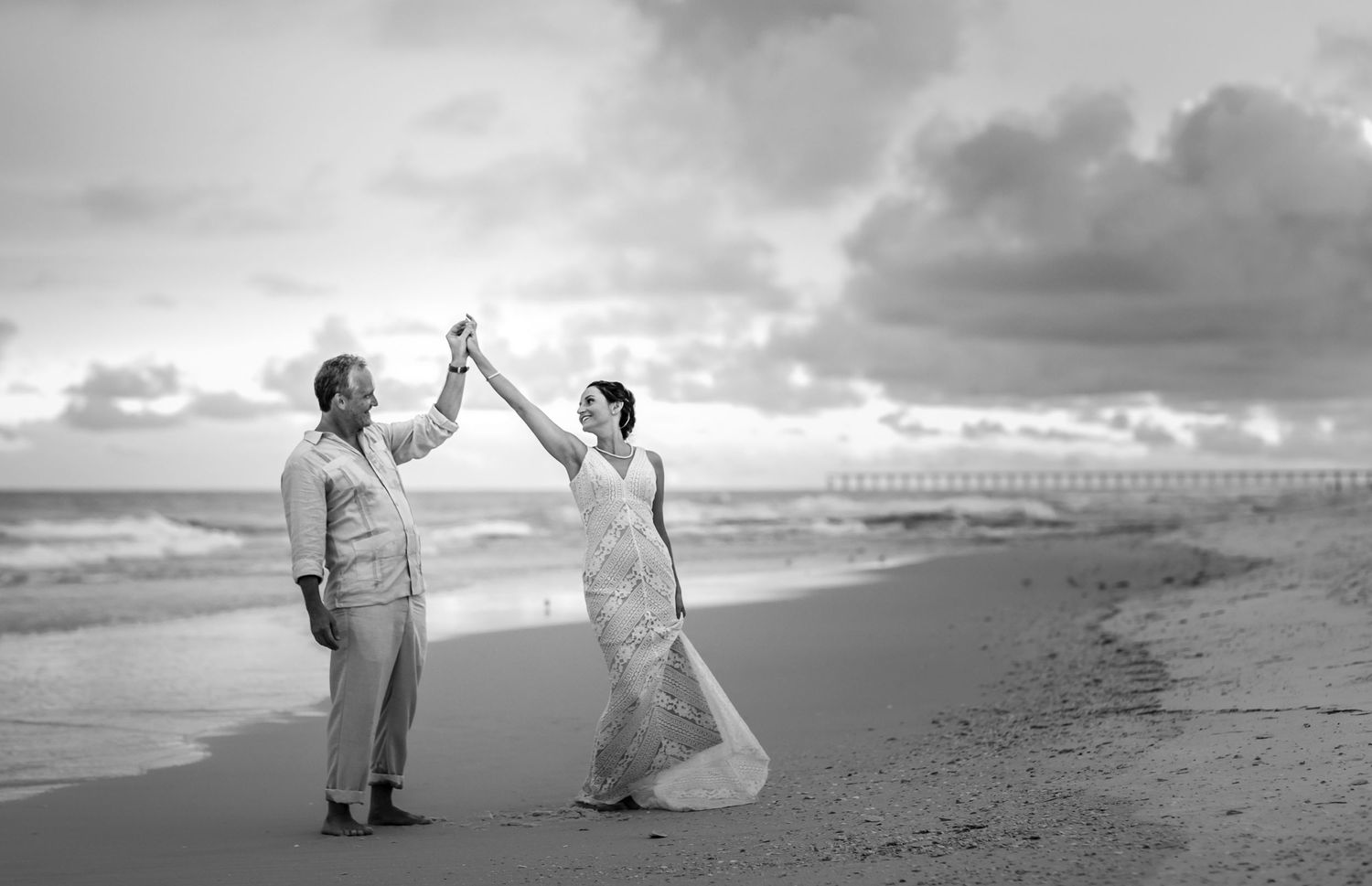 Black and white photo of couple dancing joyfully on beach in wedding attire.