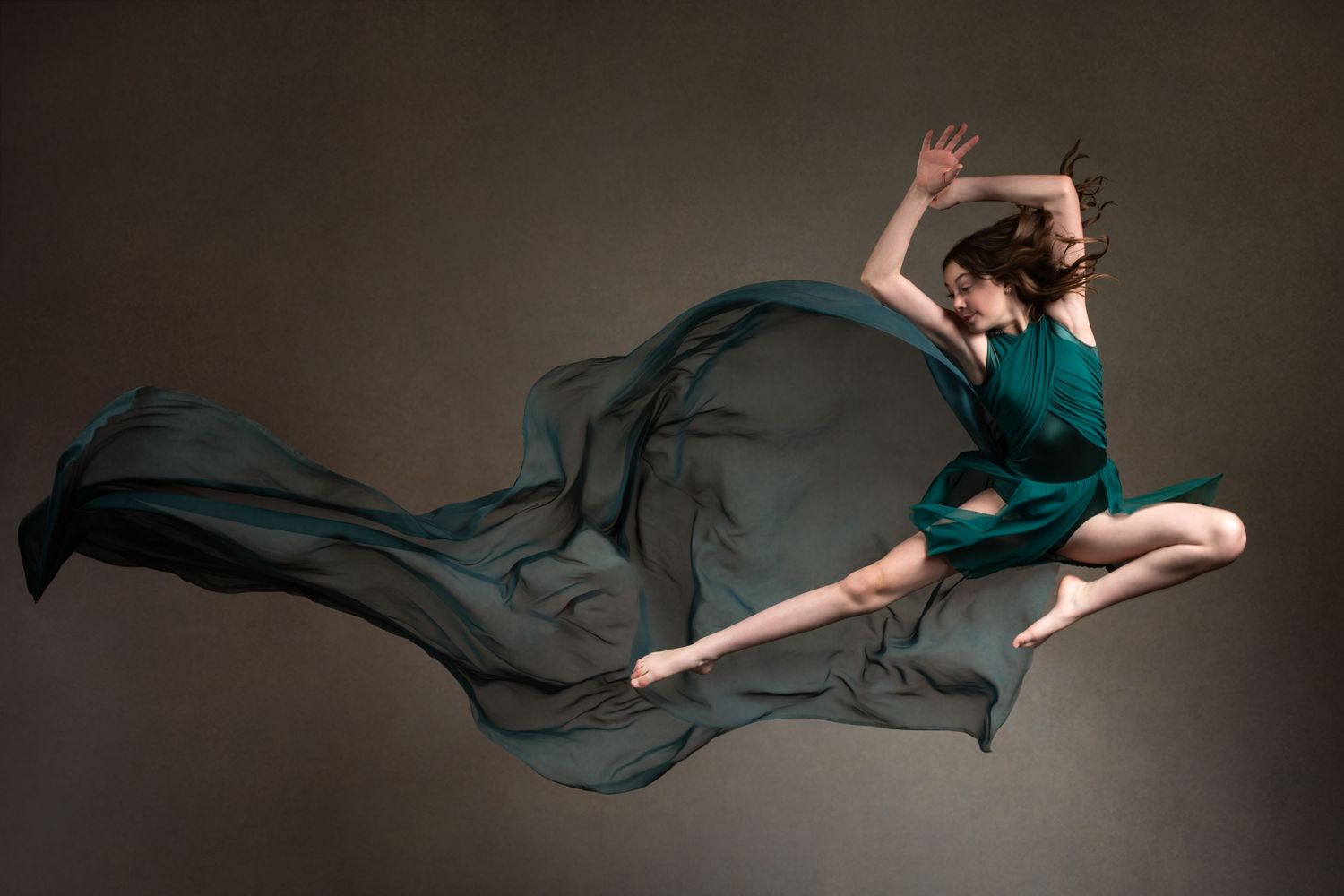 Graceful dancer in emerald dress leaping through air with flowing fabric against dark backdrop.
