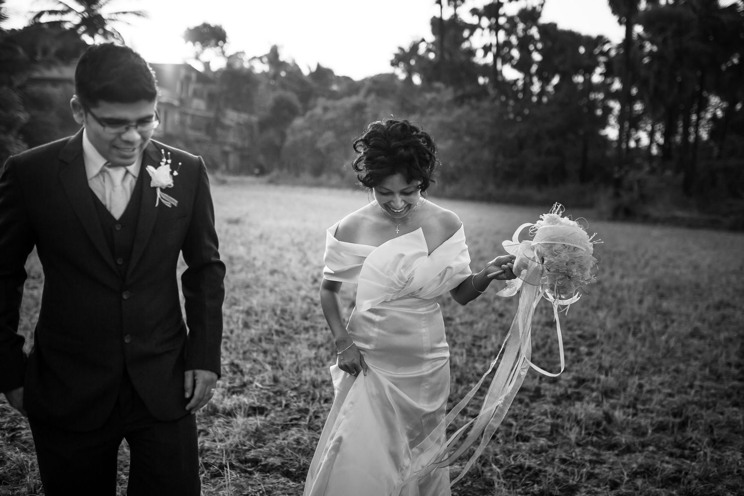 A bride and groom walk together in a field, with her holding a bouquet in a black and white photo.